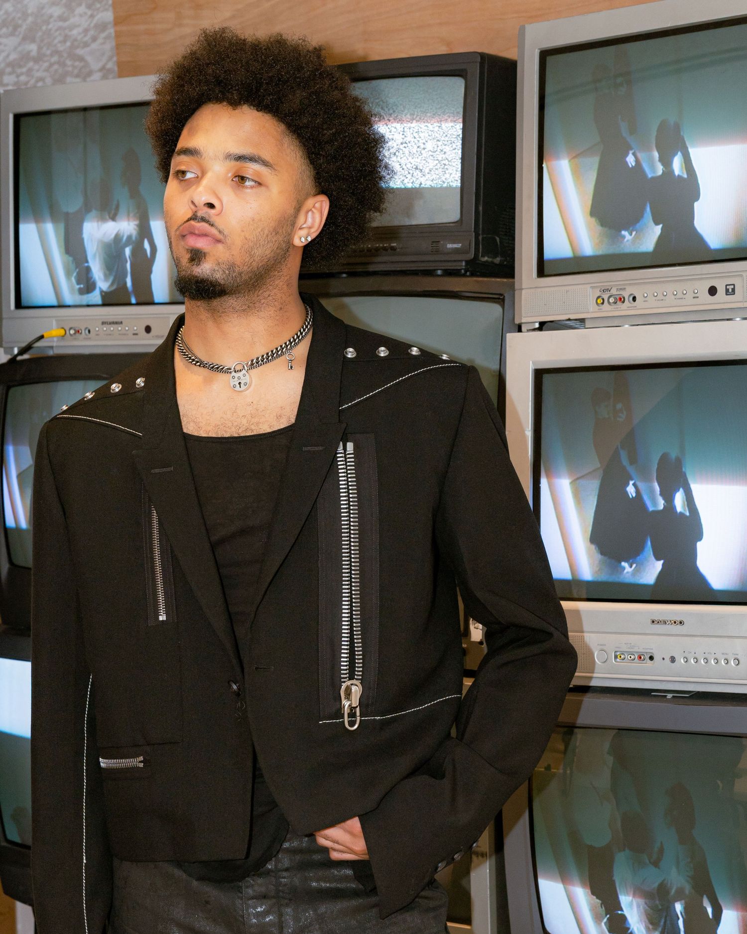 Young man with afro hair wearing black jacket poses in front of stacked vintage CRT televisions.