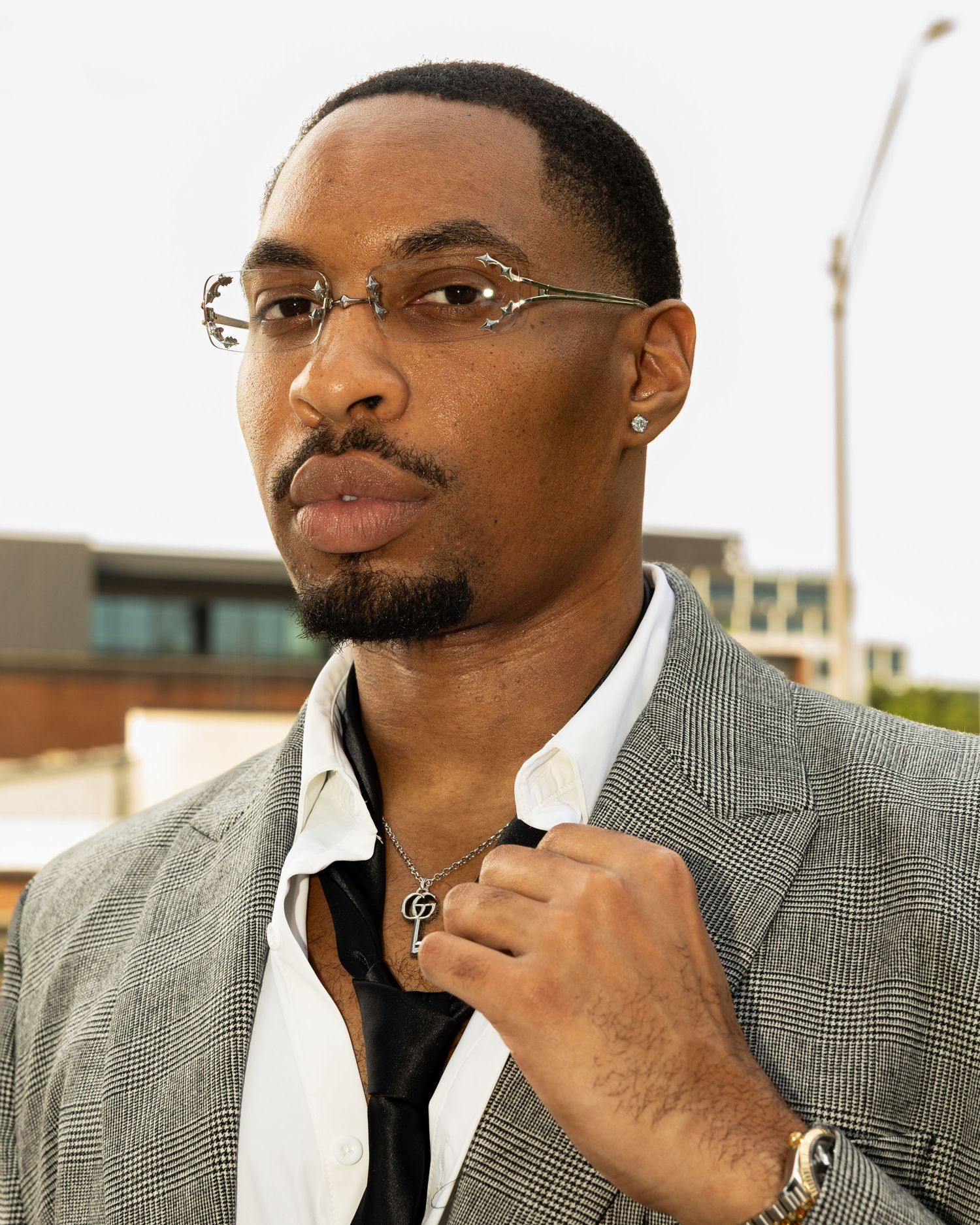 Stylish young man wearing rimless glasses and a gray blazer with a black shirt, posing outdoors.