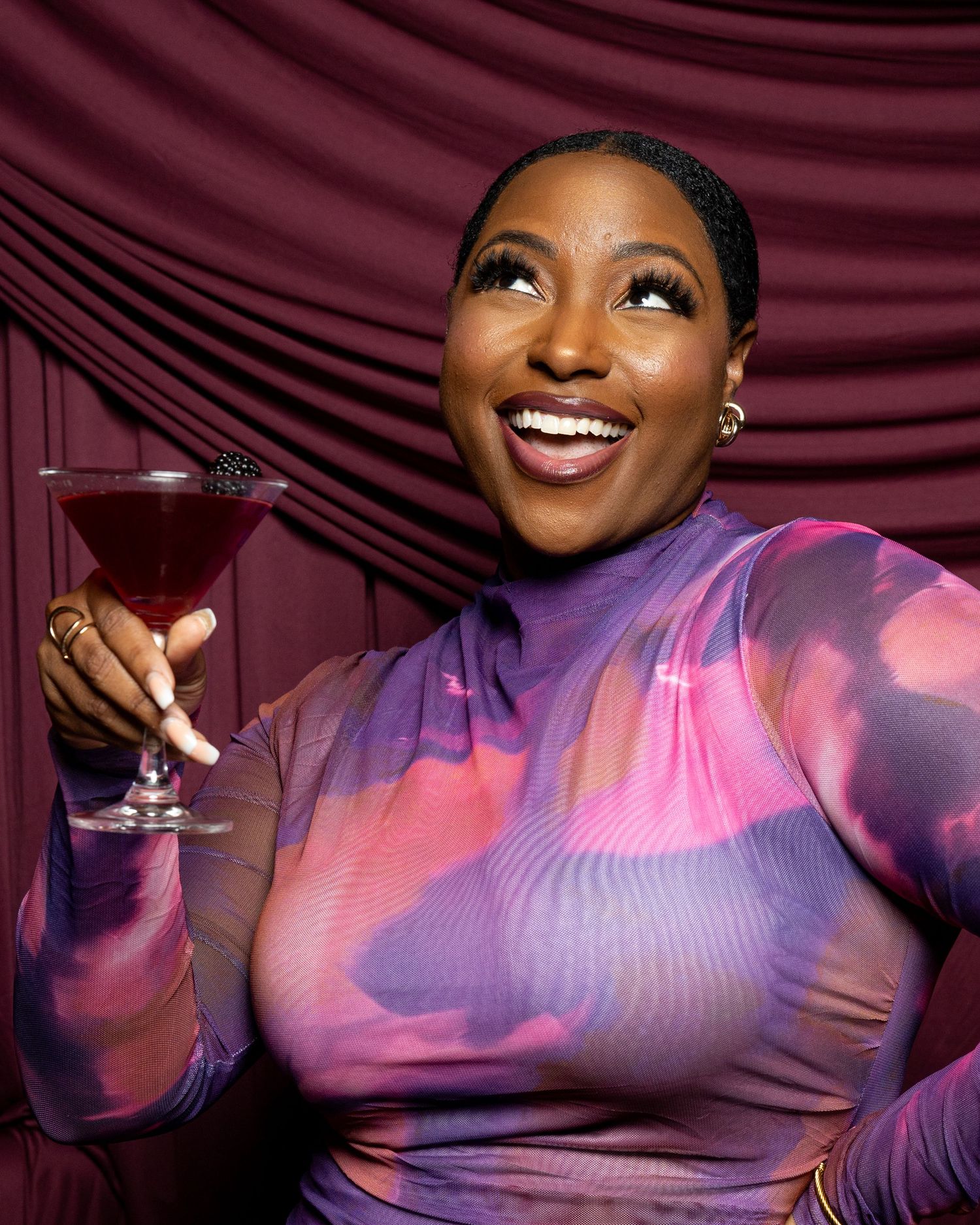 Smiling woman in colorful purple pink mesh dress holding a cocktail martini glass against burgundy draped background.