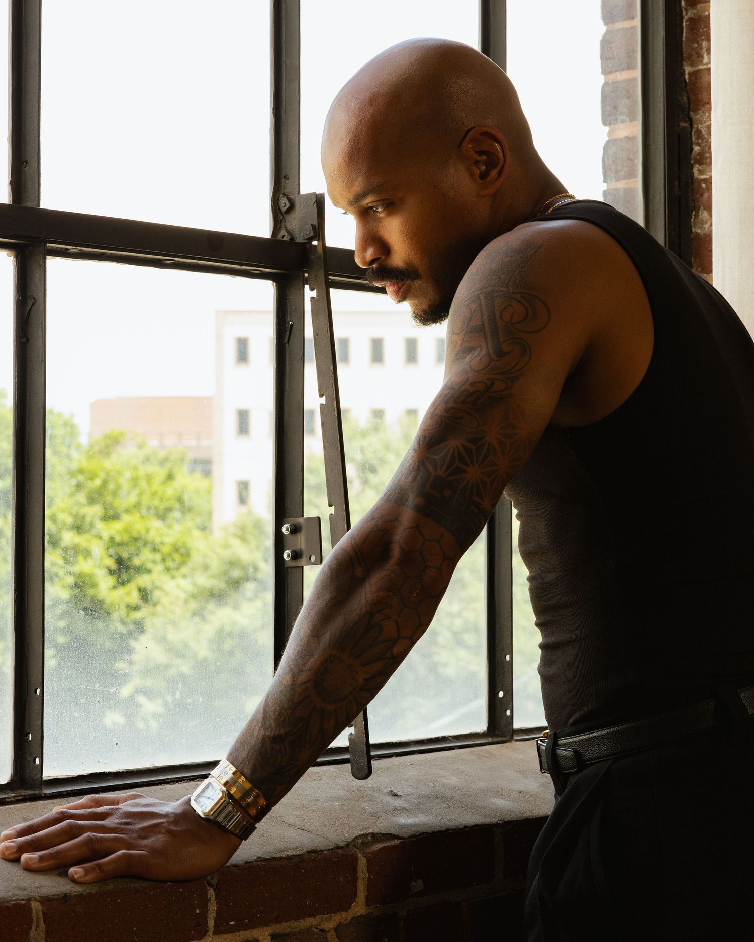 Bald tattooed man in black tank top leaning on windowsill, looking down thoughtfully with gold bracelet.