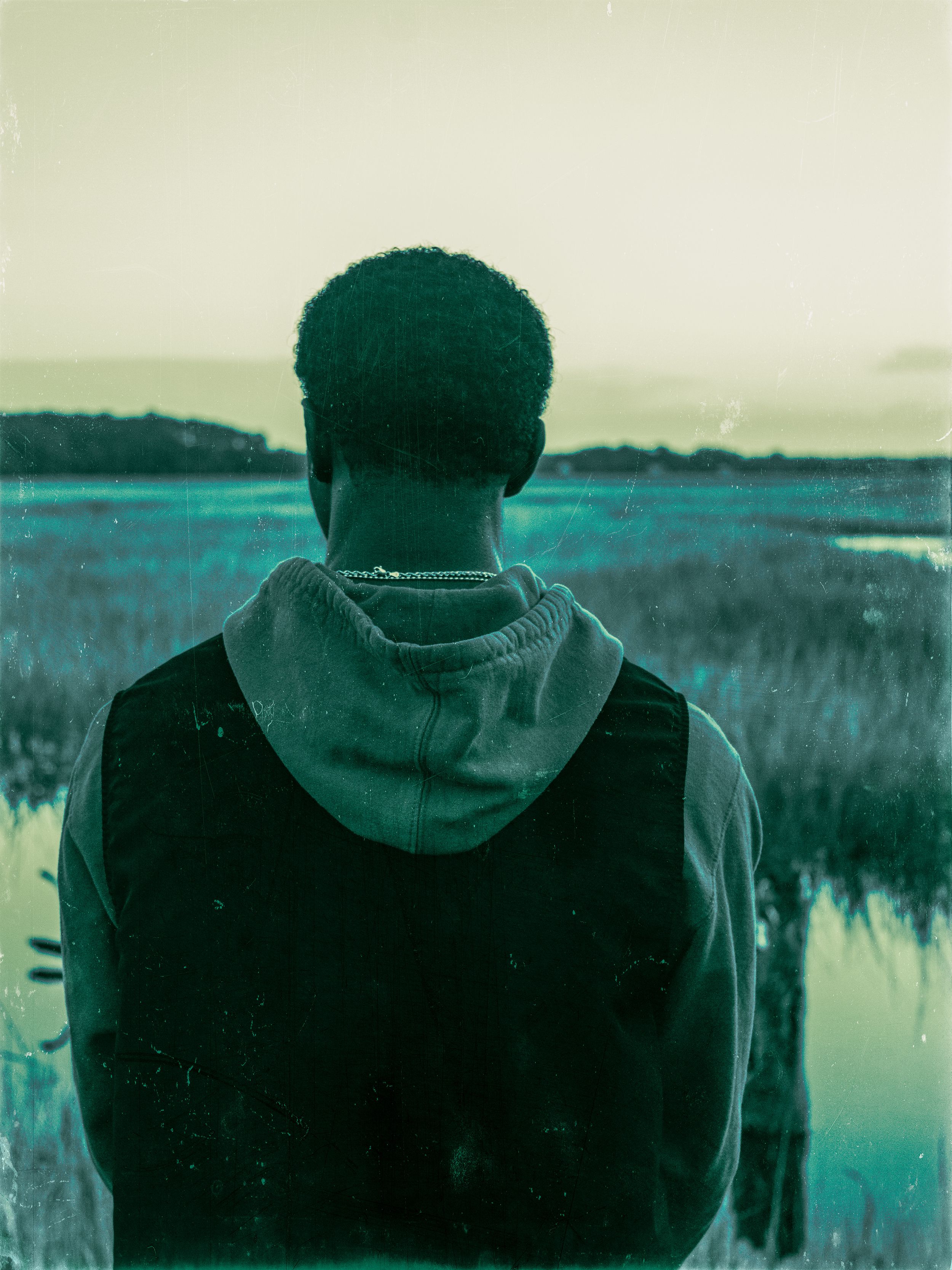 Silhouette of person in hoodie looking out over waterfront landscape at dusk.