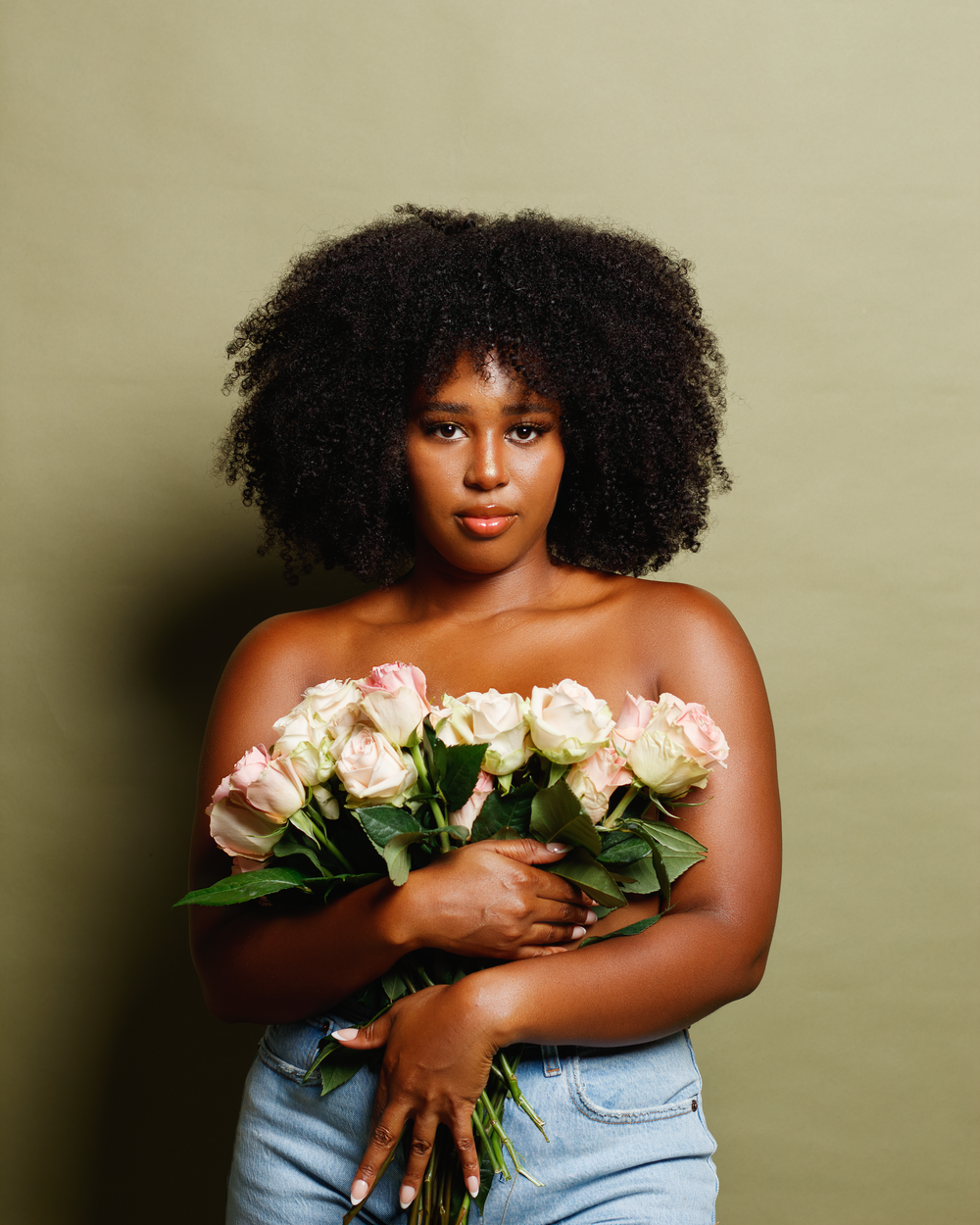 A person with natural curly hair holds a bouquet of white roses while wearing jeans against a neutral background.