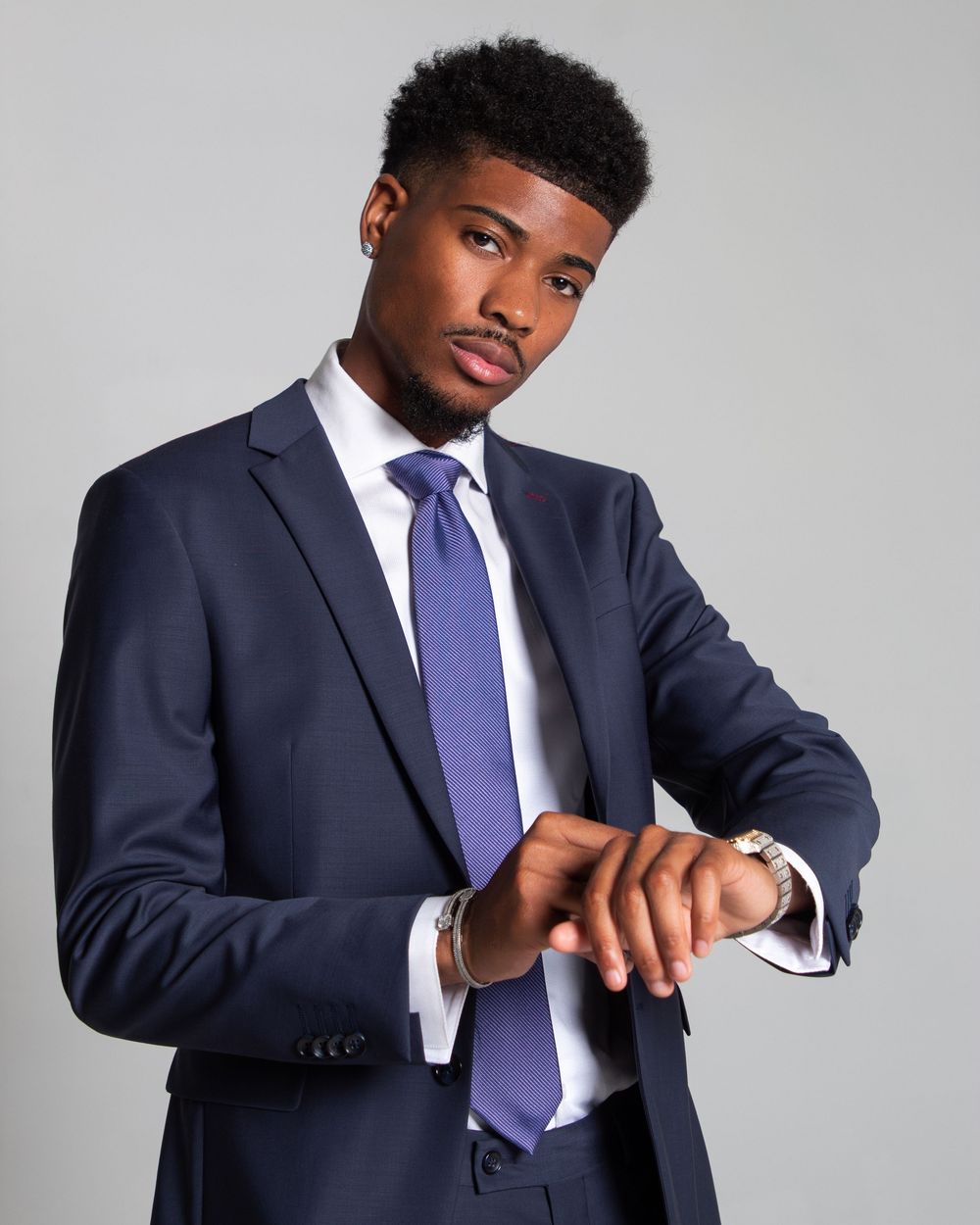 Professional wearing a navy suit and purple tie checks wristwatch while standing against a light gray background.