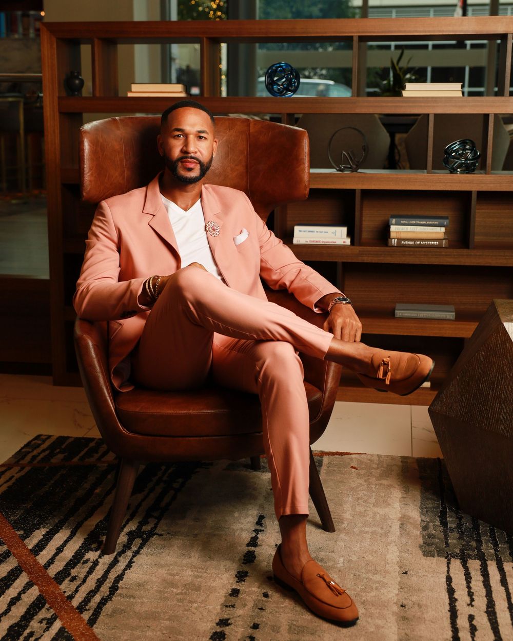 A person wearing a peach-colored suit sits confidently in a brown leather armchair against wooden shelving and patterned rug.