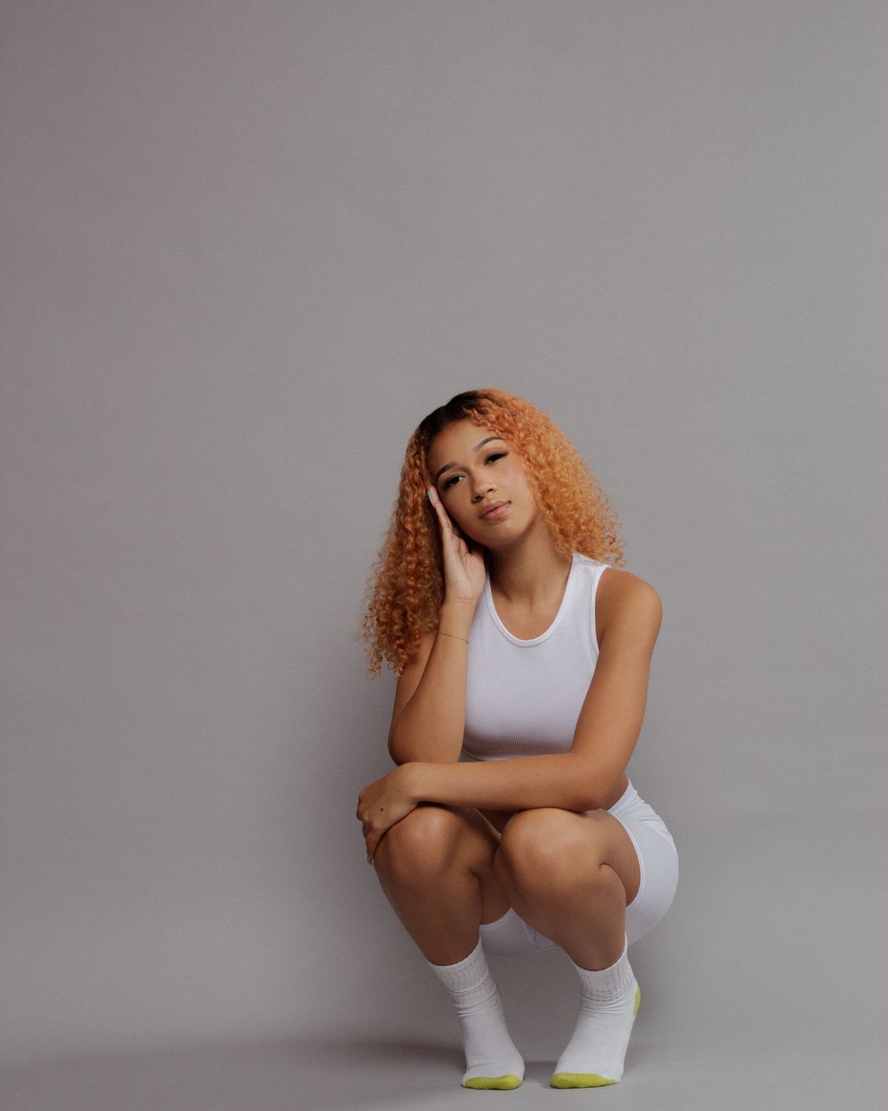 Athletic model in white tank top and shorts crouches in dynamic pose against gray backdrop with curly ginger hair.