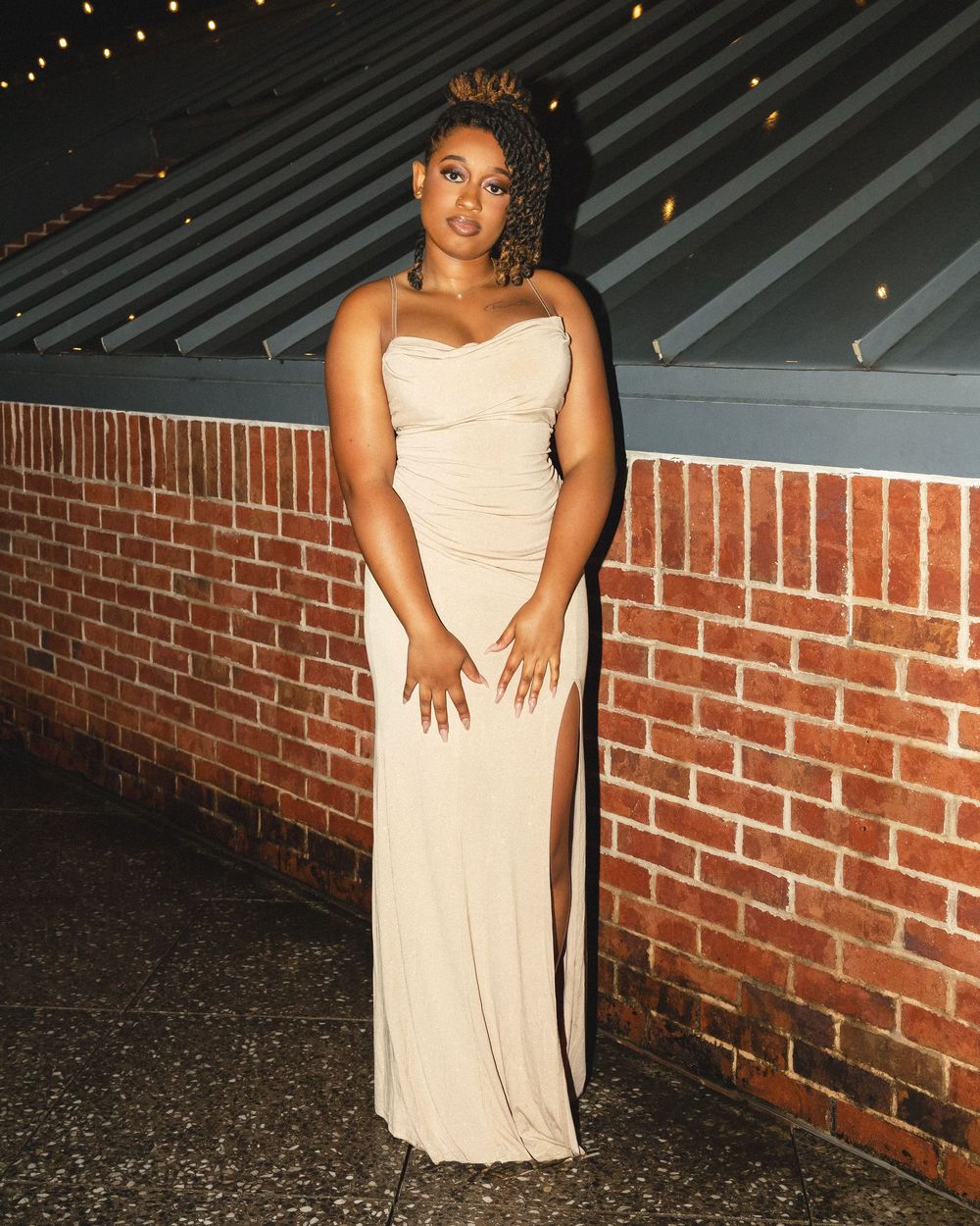 Someone in an elegant cream-colored strapless gown poses against a brick wall under string lights at night.