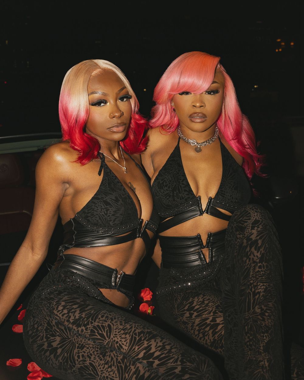 Two women in matching black outfits with bright pink hair pose together against a dark background.