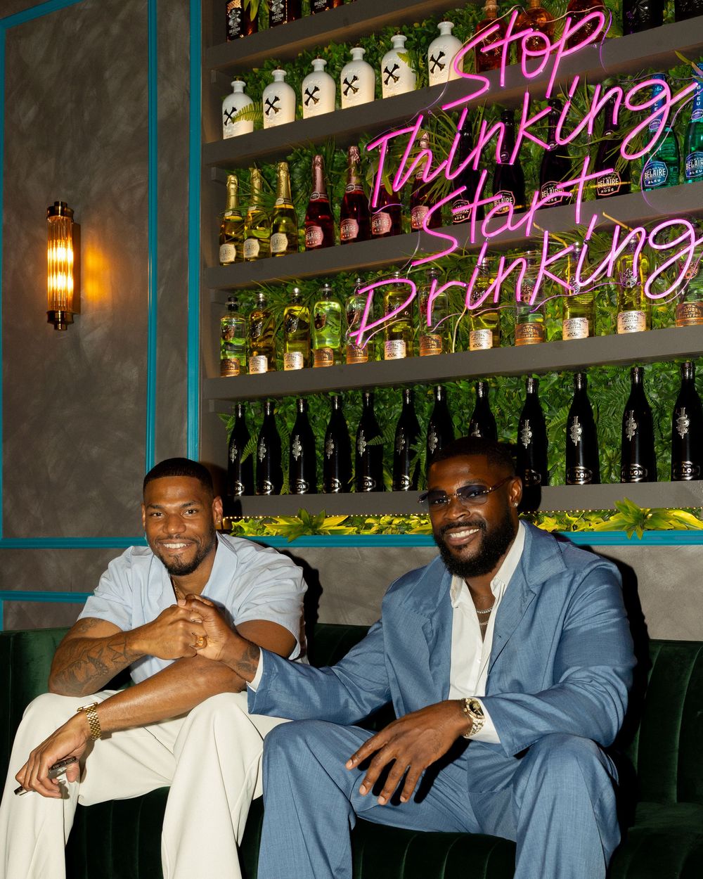Group of men in blue suits sitting casually at a bar with illuminated green bottles displayed on shelves behind them.