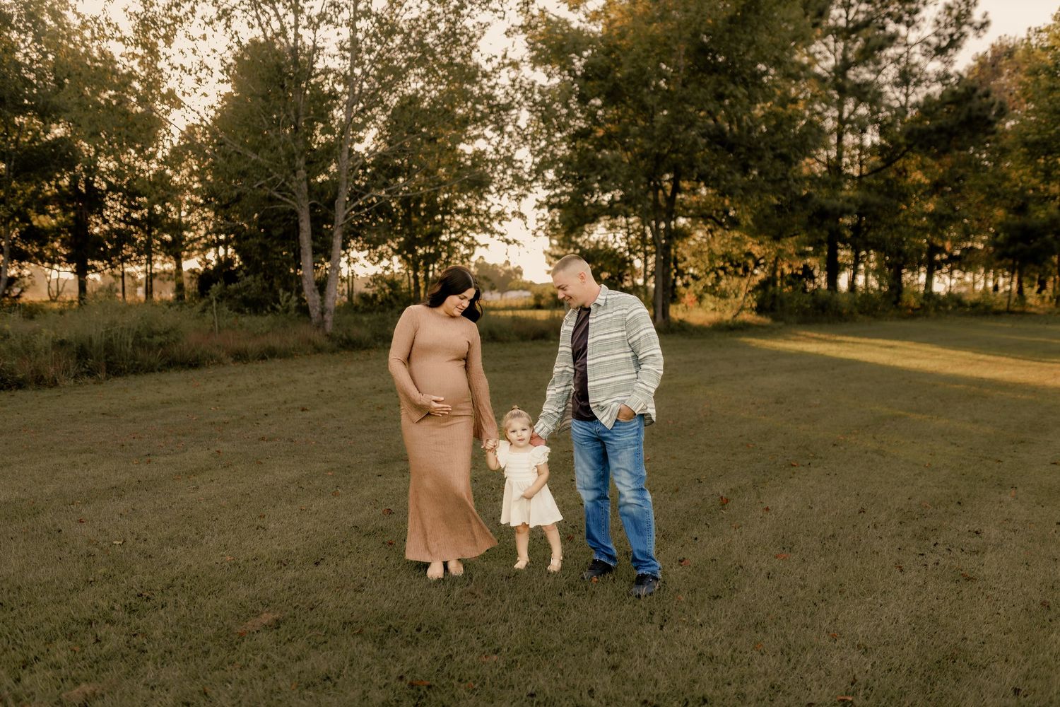 Family of three holds hands while walking in field during sunset photo session.