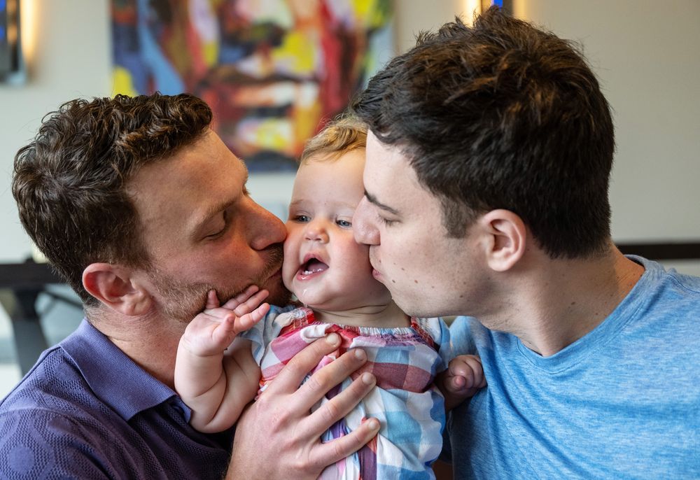 Two men kissing a laughing baby on the cheeks, sharing a joyful family moment indoors.