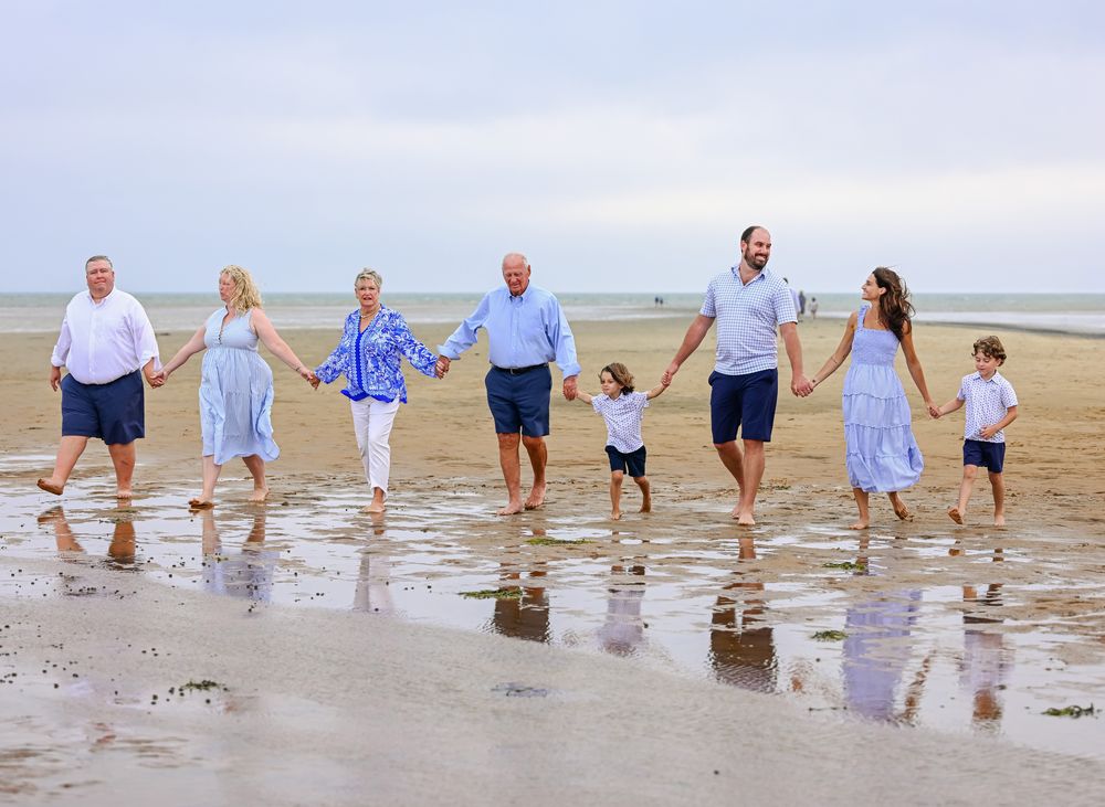 Multi-generational family holding hands walking on a sandy beach on a cloudy day in coordinating blue and white outfits.