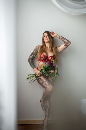 Artistic boudoir photo series showing a woman in red lingerie with tattoos holding roses against a white wall.