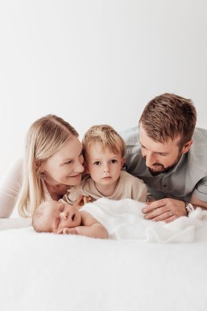 A family lies together on white bedding while gazing lovingly at a newborn baby.