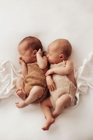 Two sleeping newborn babies wearing knit rompers lie together on white fabric in a sweet pose.