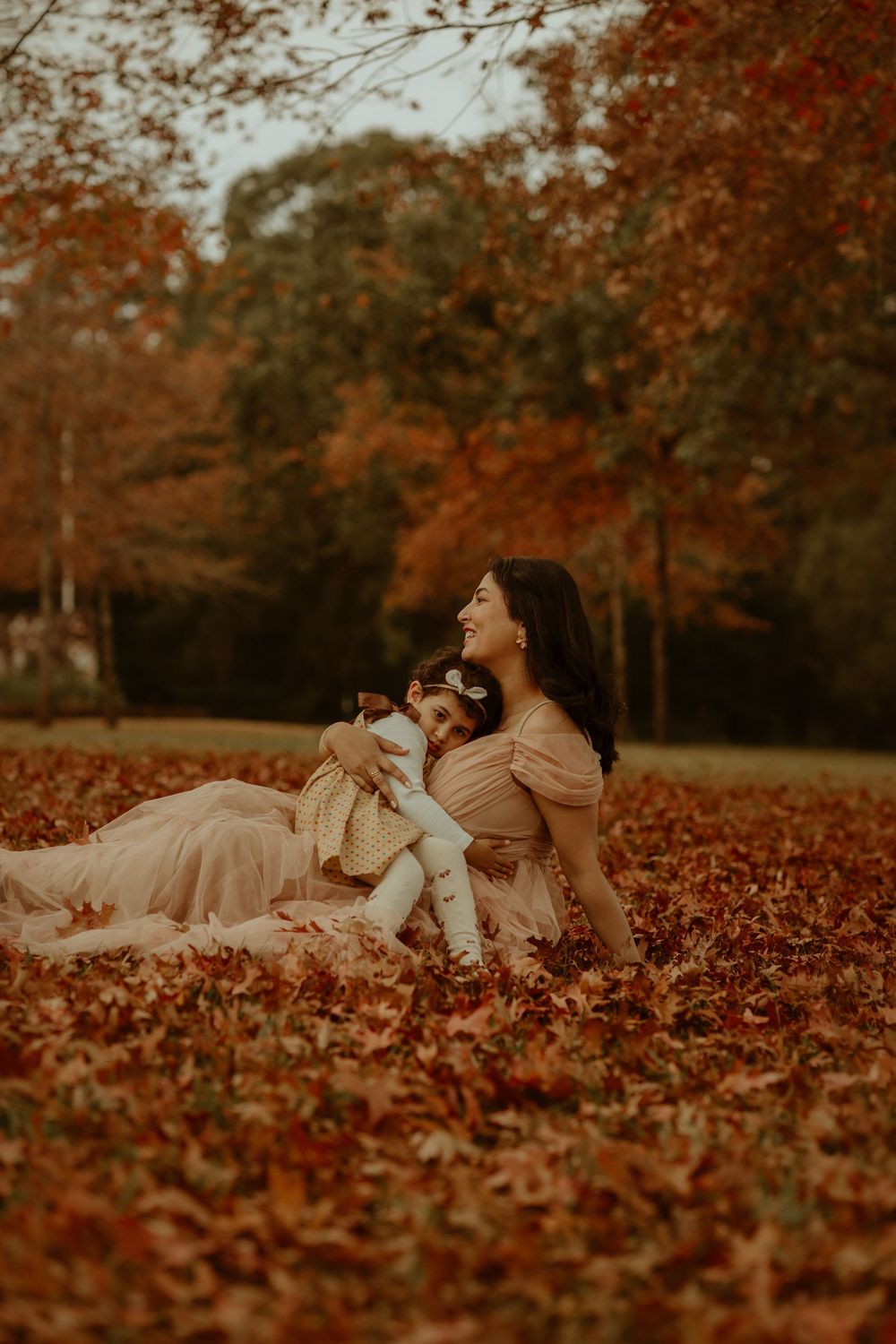 Mother and baby girl sitting in autumn leaves wearing pink dresses, laughing together in a park with red fall foliage.