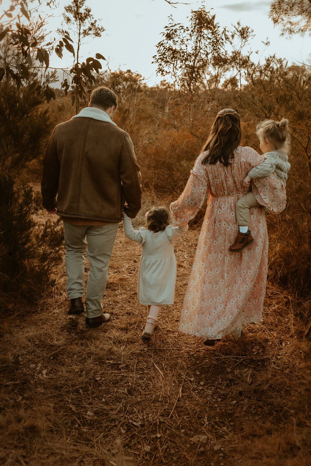 A family walks together on a hillside path in sepia tones, holding hands and wearing flowing dresses.