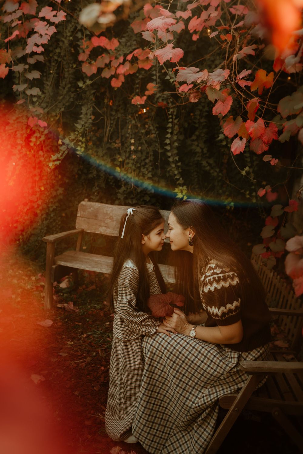 A mother and daughter share a tender nose-to-nose moment on a garden bench surrounded by vibrant autumn foliage.