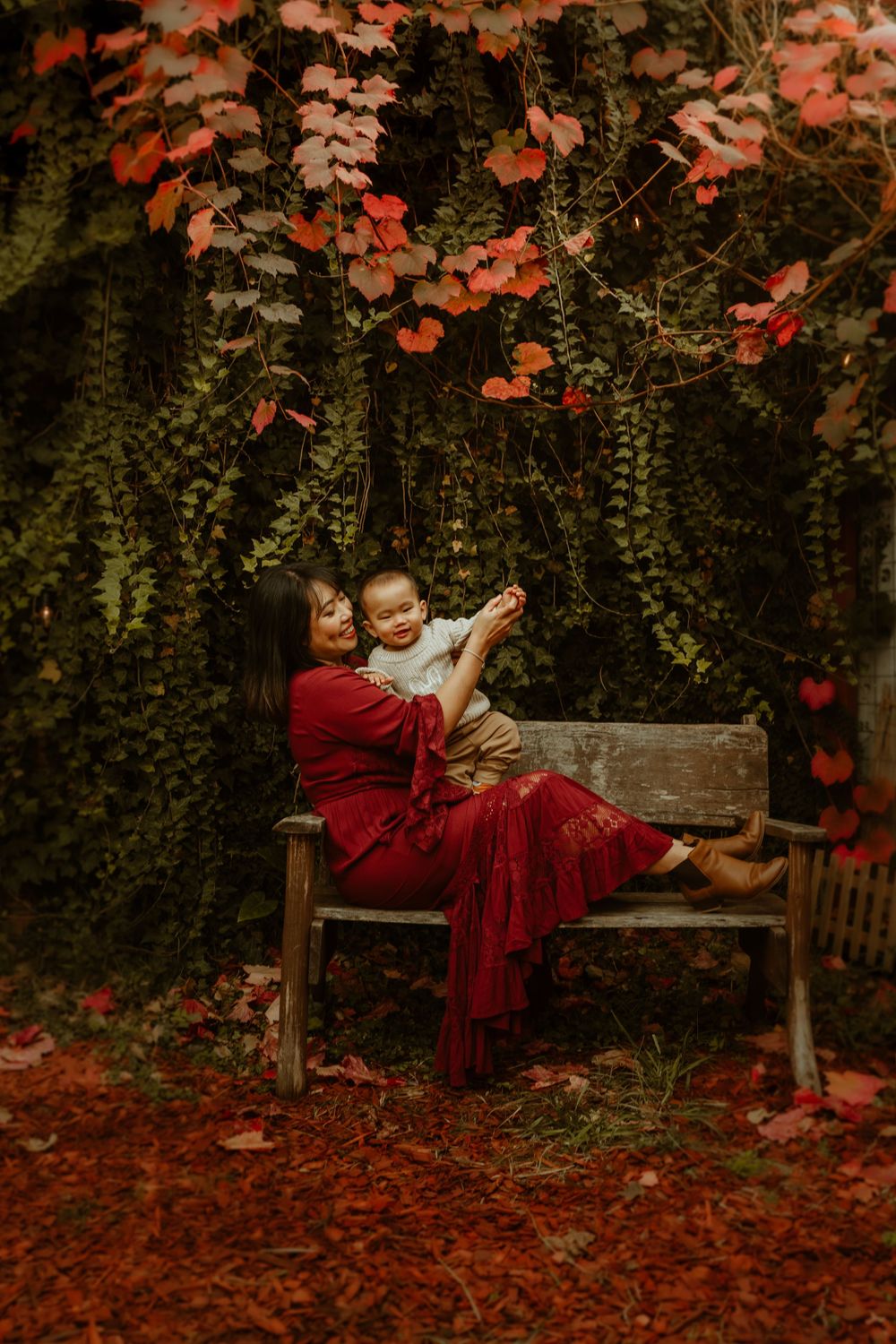 Mother in red dress sits on bench holding baby amid autumn foliage and fallen leaves.