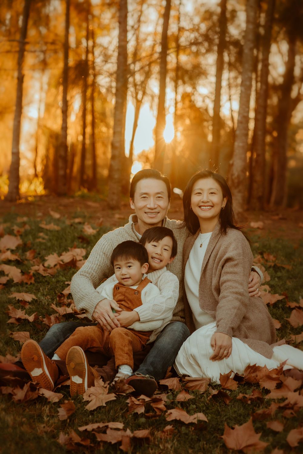 Family of four sitting on autumn leaves at golden hour sunset in a forest park, smiling warmly at camera.
