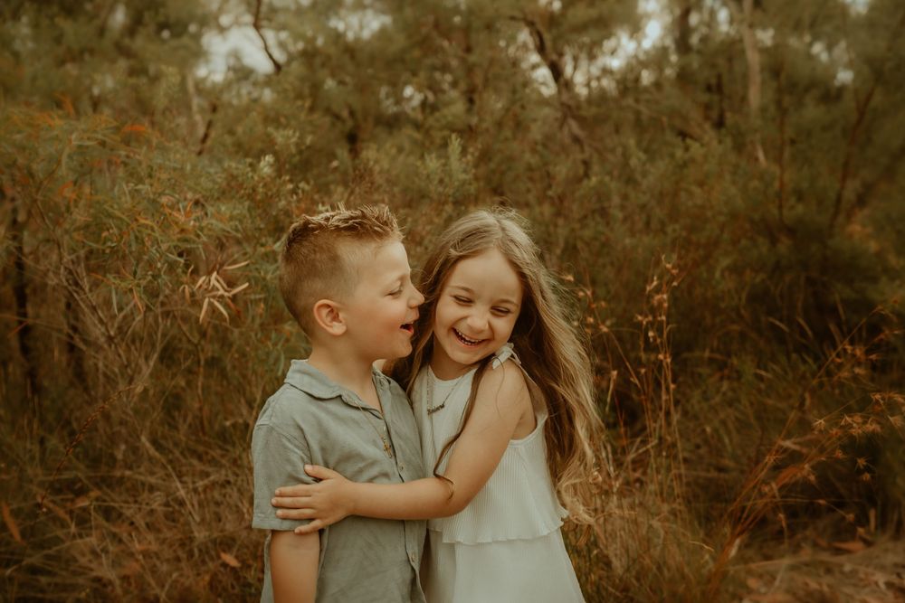 Two young siblings laughing and hugging outdoors in a natural bush setting, captured in warm earthy tones.