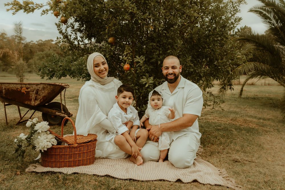 Muslim family of four smiling together on a picnic blanket under a fruit tree in a lush garden setting.