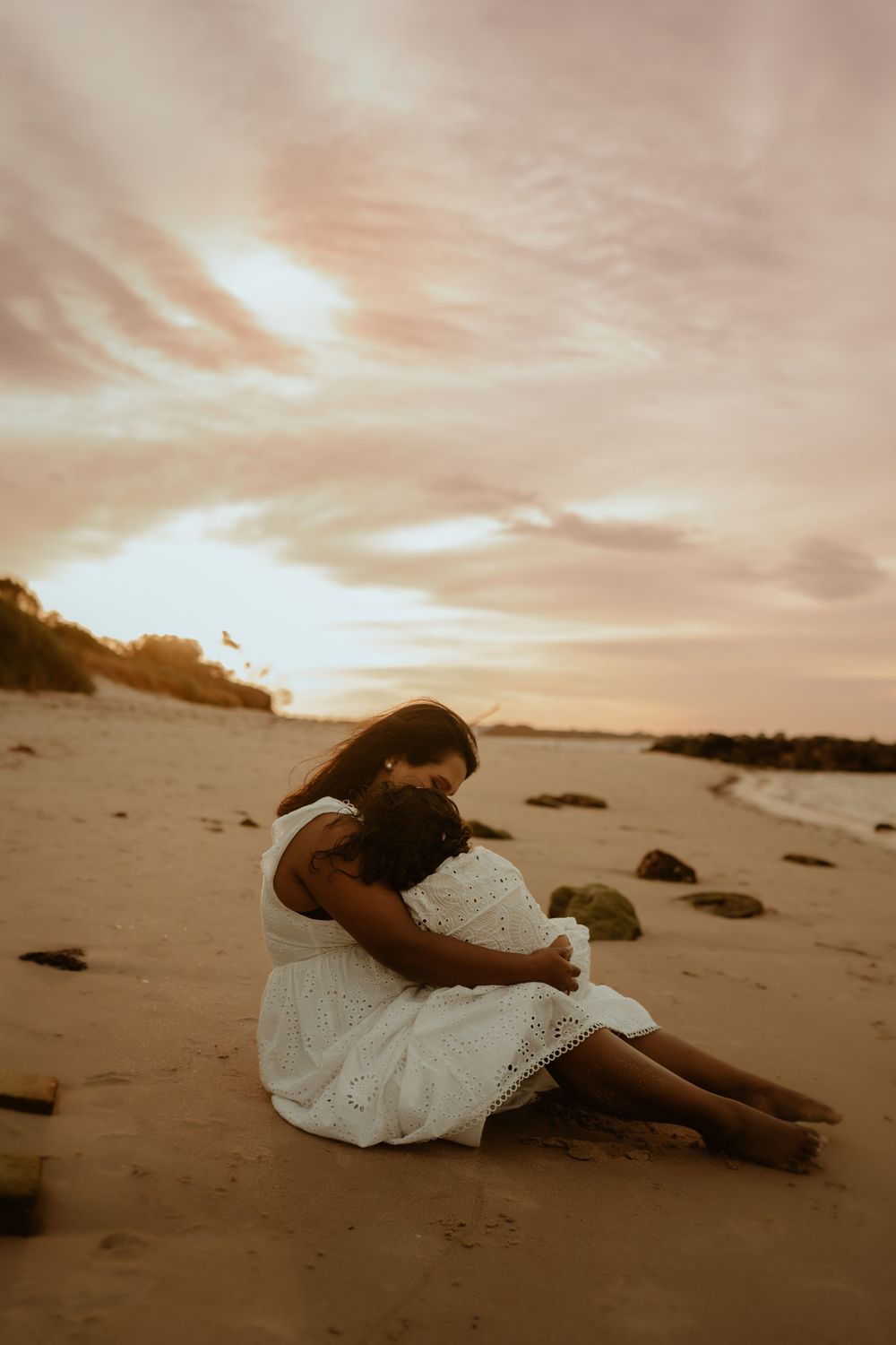 A figure in a white dress reclines on a sandy beach at sunset creating a dreamy, artistic silhouette.