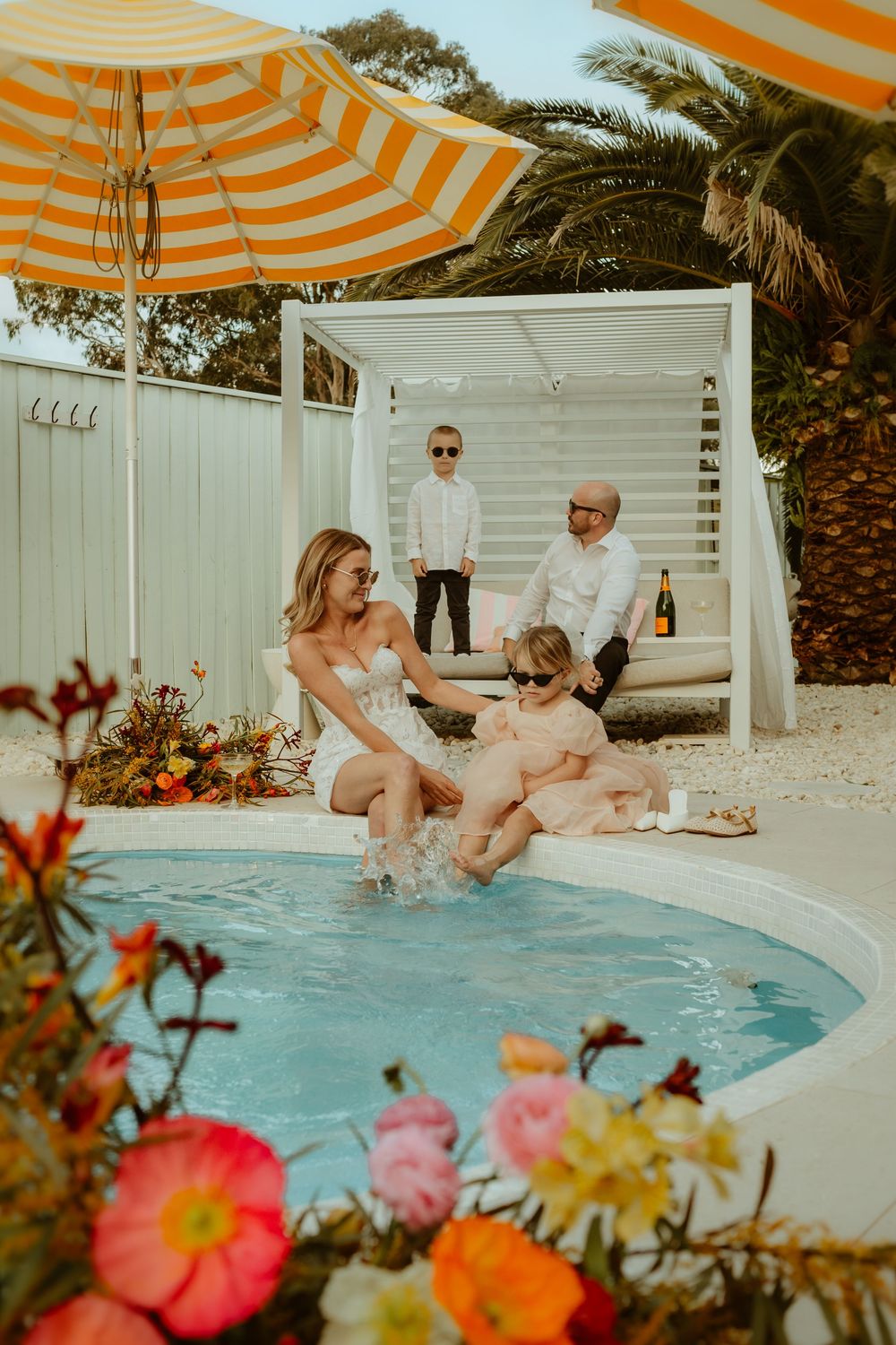 A family enjoys a summer pool day with orange striped umbrellas and colorful flowers surrounding a backyard swimming pool.