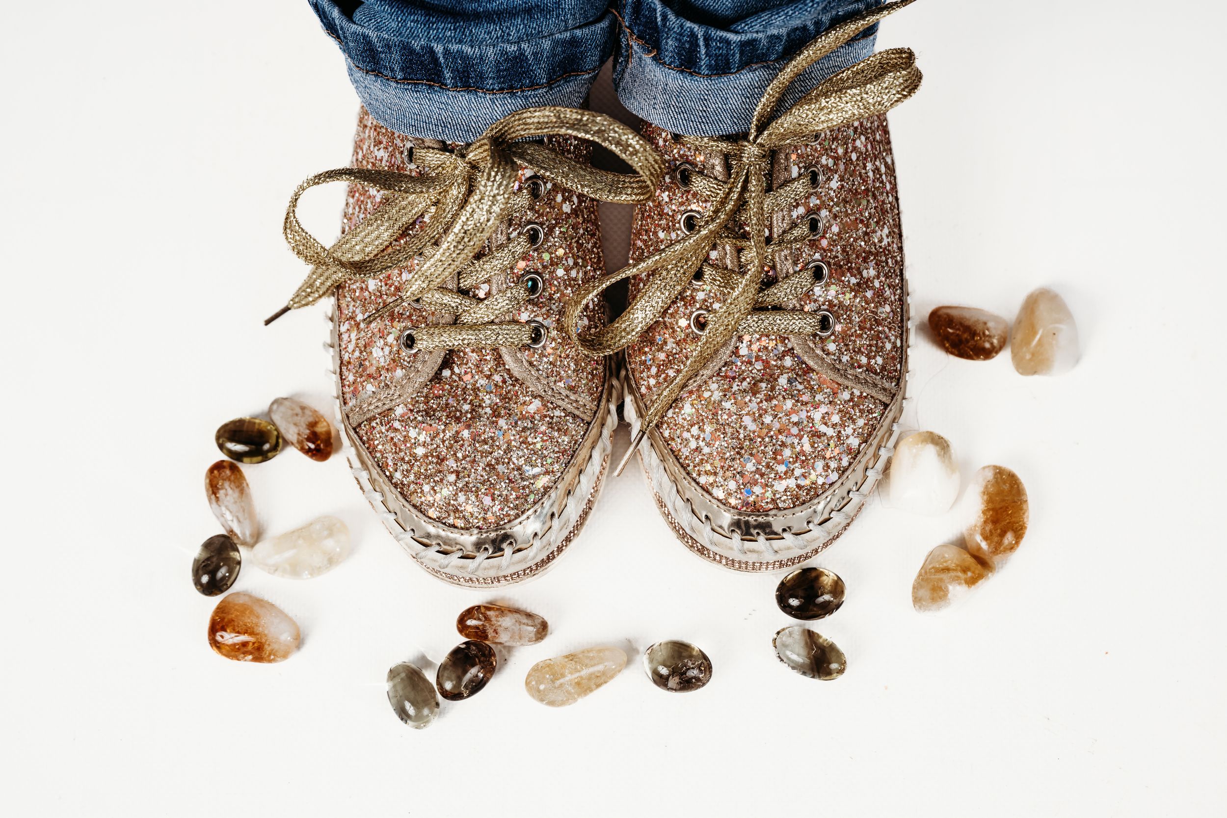Rose gold glitter sneakers with gold laces surrounded by decorative stones on white background.