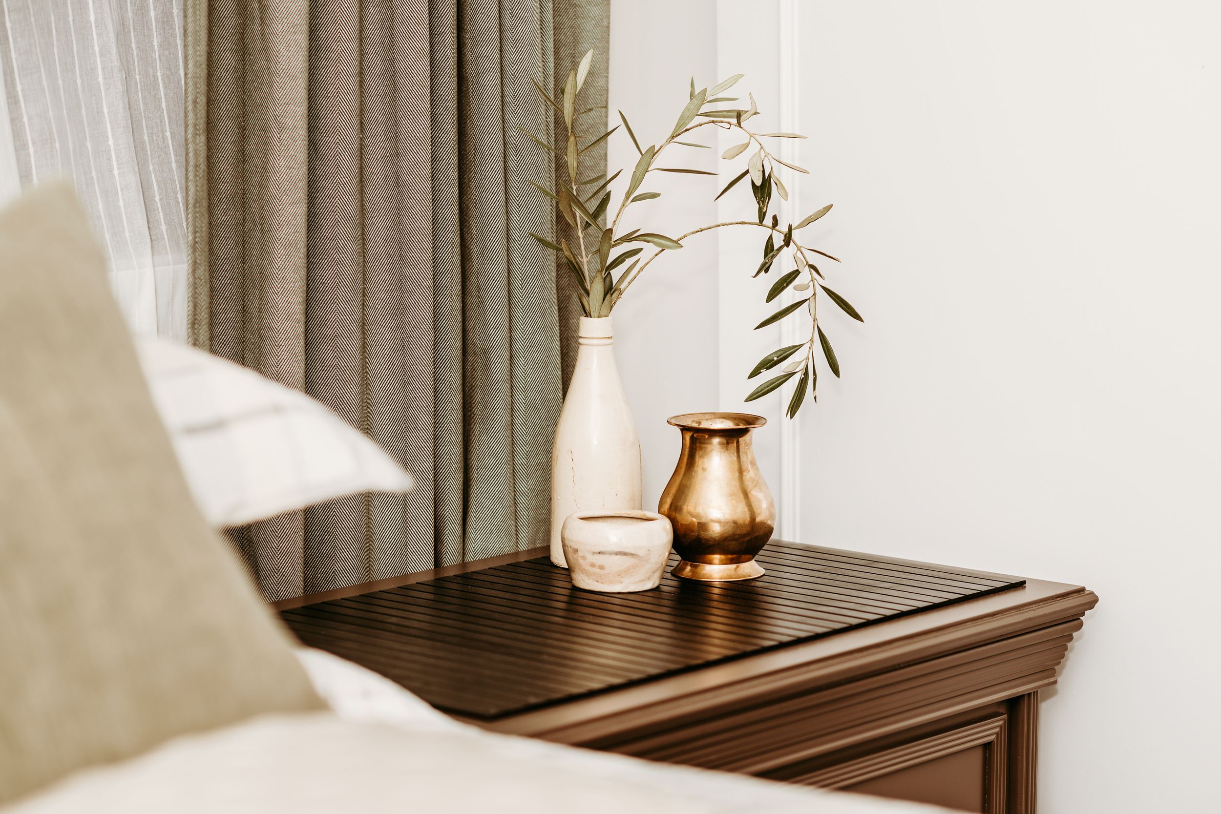 A copper vase with olive branches sits atop a dark wooden nightstand next to beige curtains and throw pillows.