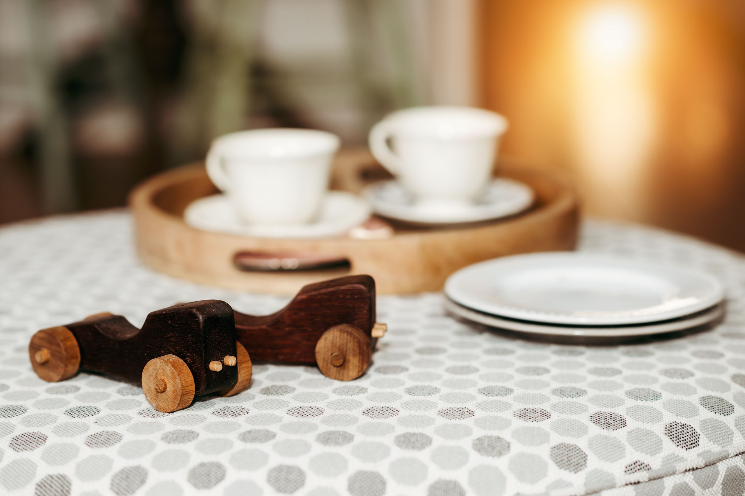 Wooden toy car sits on lace tablecloth next to white coffee cups and saucers with plates in background.