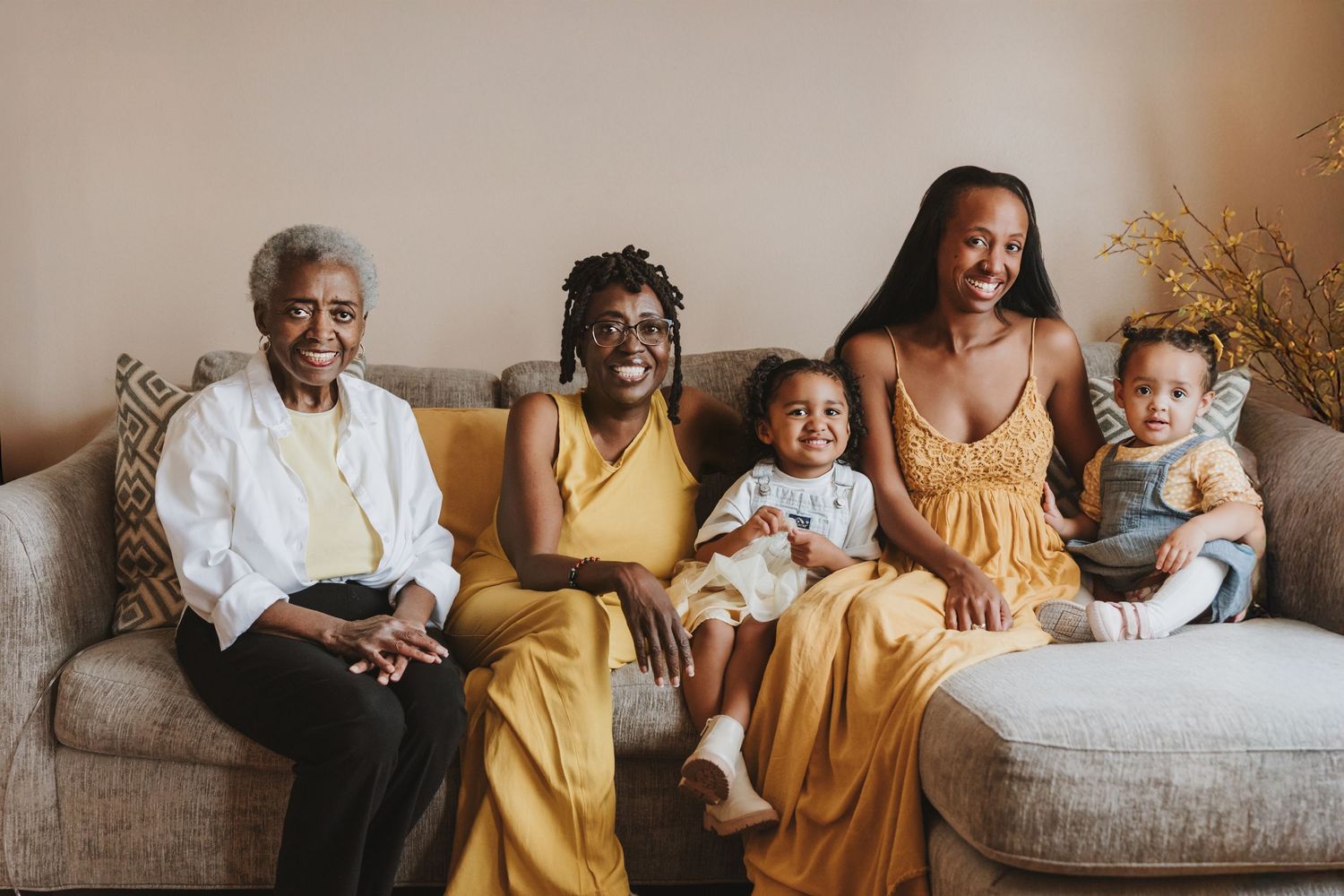 Four generations of a family wearing yellow and white sit together and smile on a beige couch.