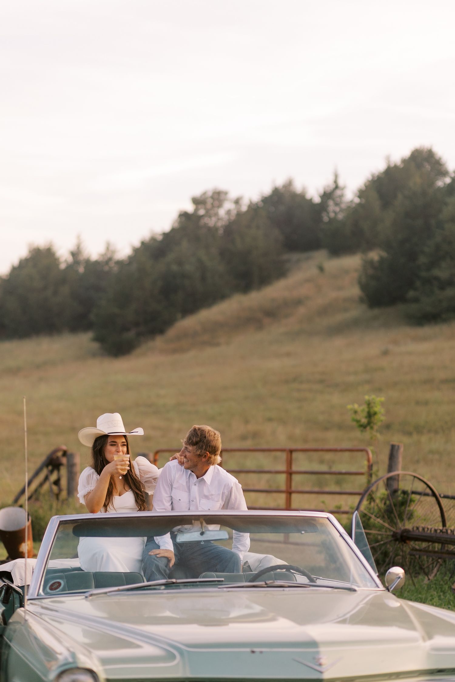 A couple sits in a vintage convertible car parked near a rustic fence in a rural setting.