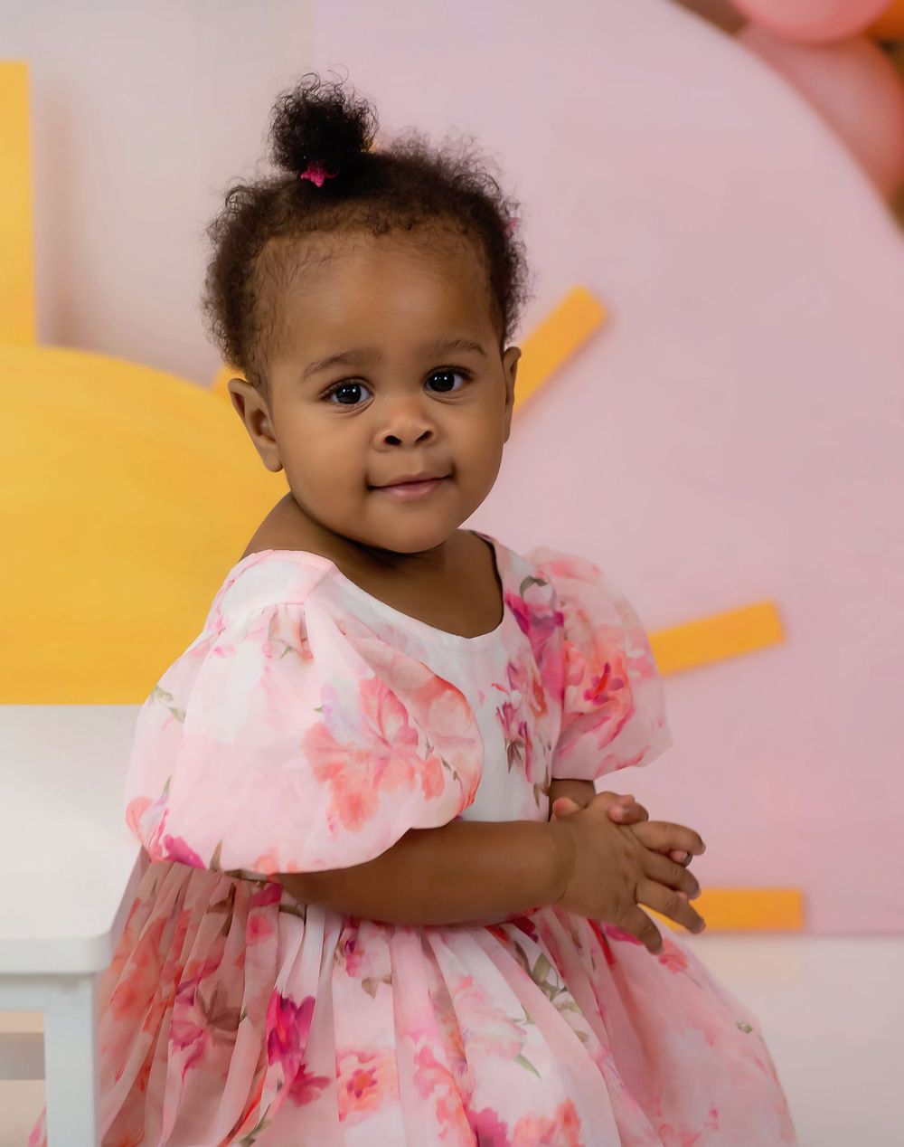 Adorable toddler girl in a pink floral dress with puff sleeves, posing sweetly against a colorful pastel background.