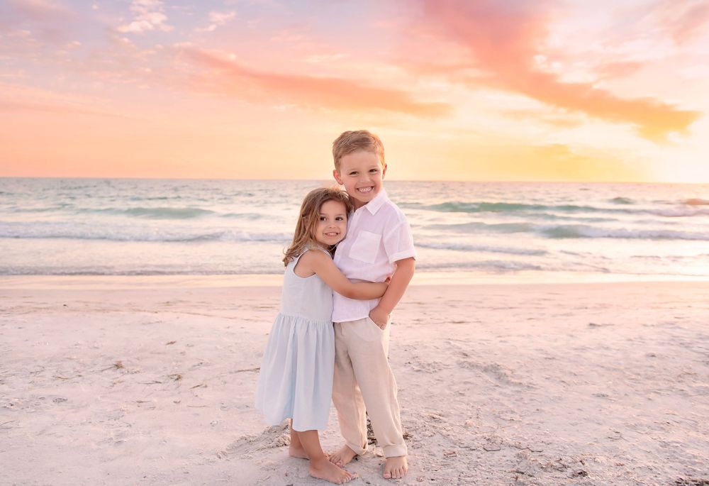 Two children embrace on a sandy beach at sunset with pastel pink and orange skies over calm turquoise waters.