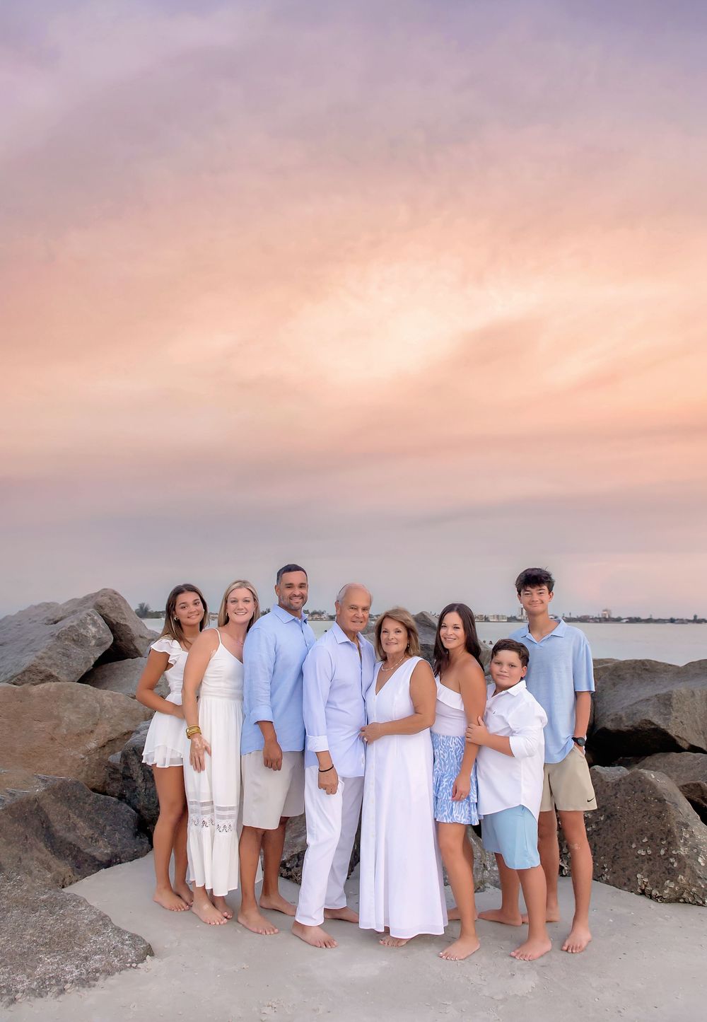 Family of eight poses on rocky beach at sunset with pastel pink and blue sky backdrop, wearing white and light blue outfits.