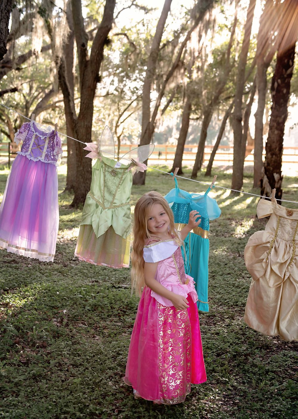 Young girl in pink princess dress stands outdoors among colorful Disney princess costumes hanging on a clothesline.