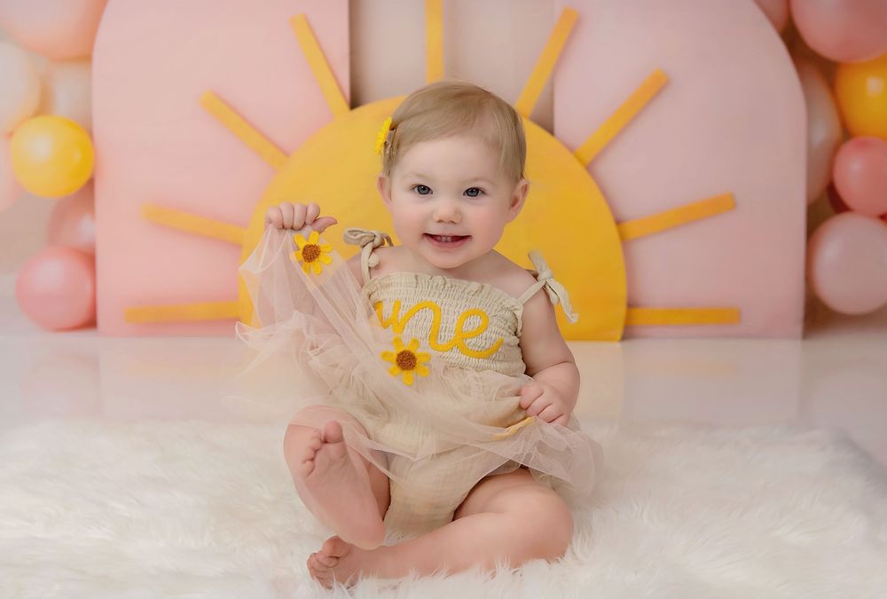 Baby girl in a tulle romper with sunflowers sits smiling at a sunshine-themed first birthday cake smash photo session.