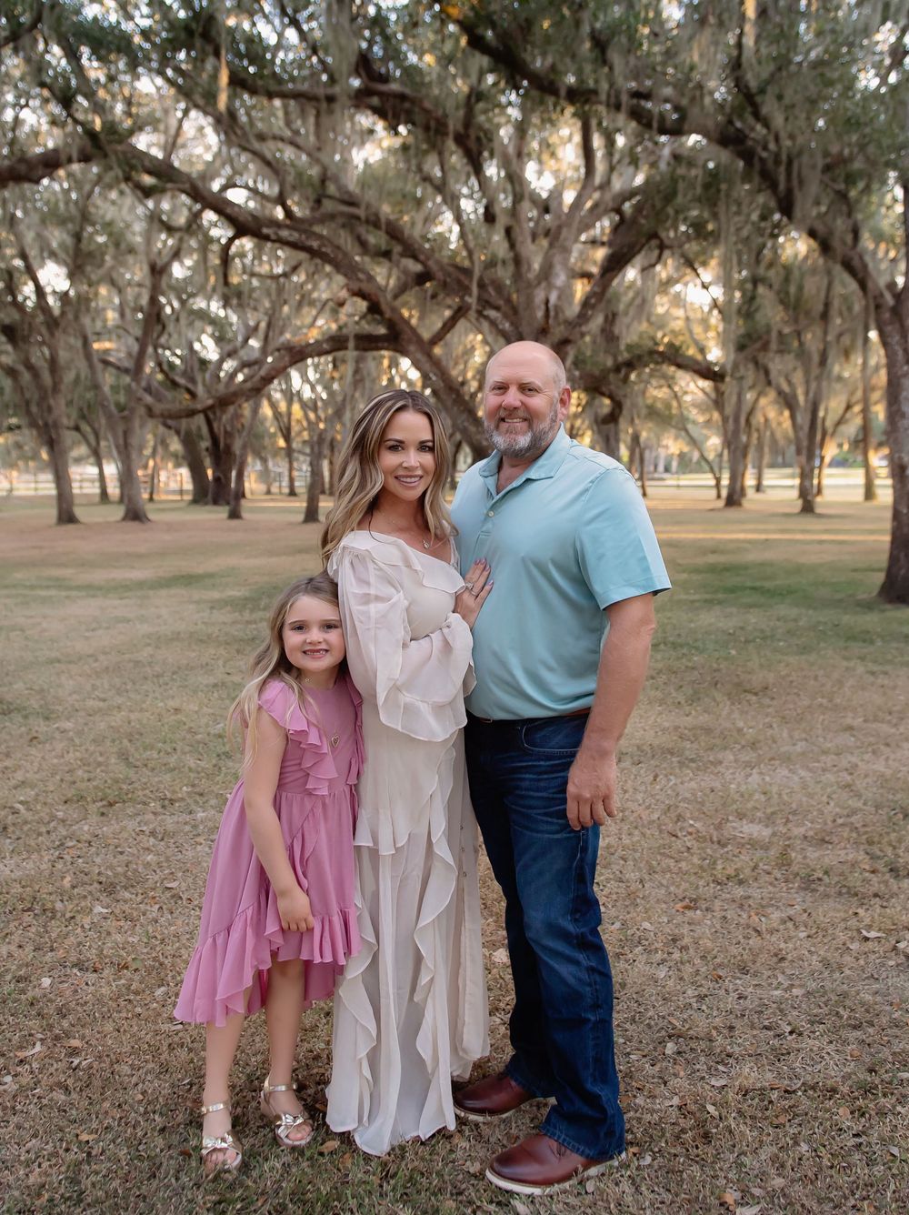 A family of three poses together outdoors under moss-draped oak trees at sunset.