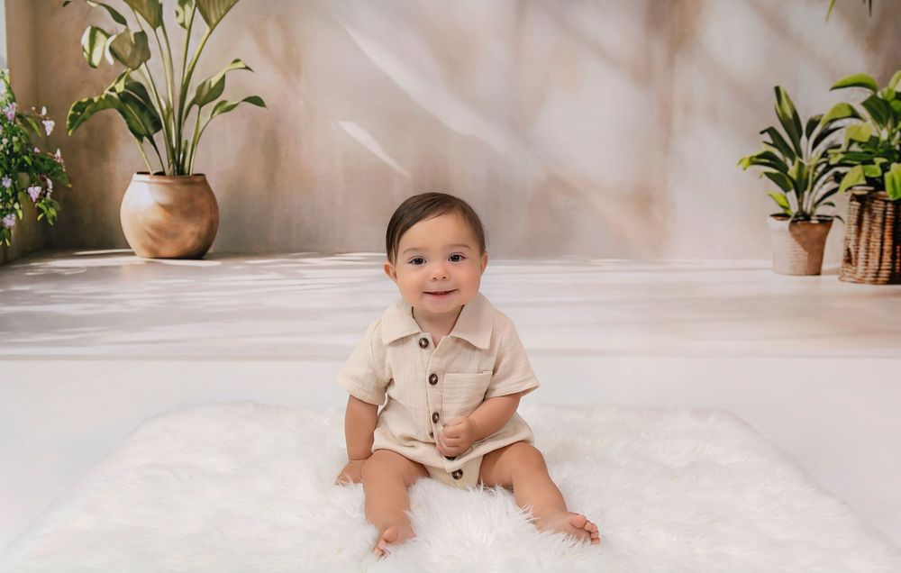 Smiling baby boy in beige romper sitting on fluffy white rug surrounded by tropical plants in bright studio setting.
