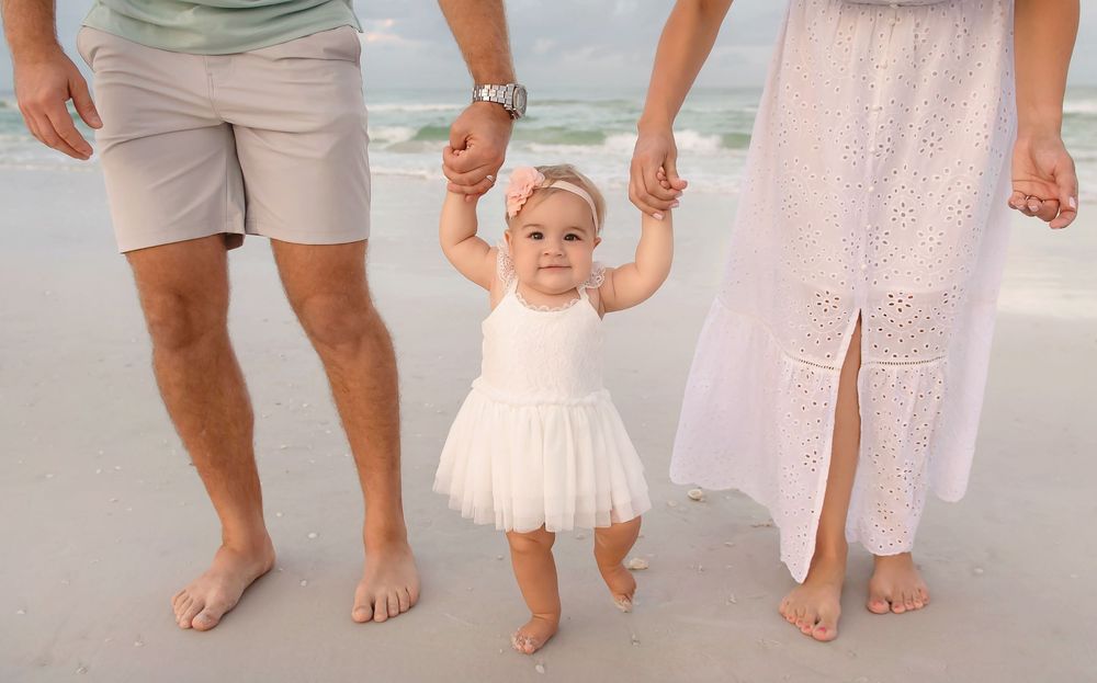 A baby girl in a white tutu dress takes first steps on a sandy beach, held by parents in matching white and tan outfits.
