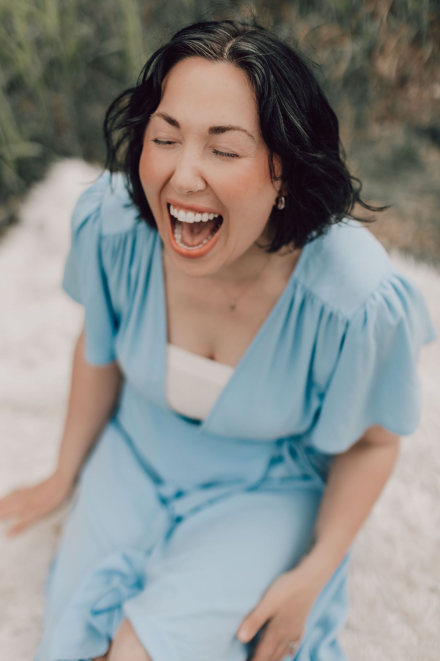 A woman in a light blue wrap dress laughing joyfully while sitting outdoors on a path.