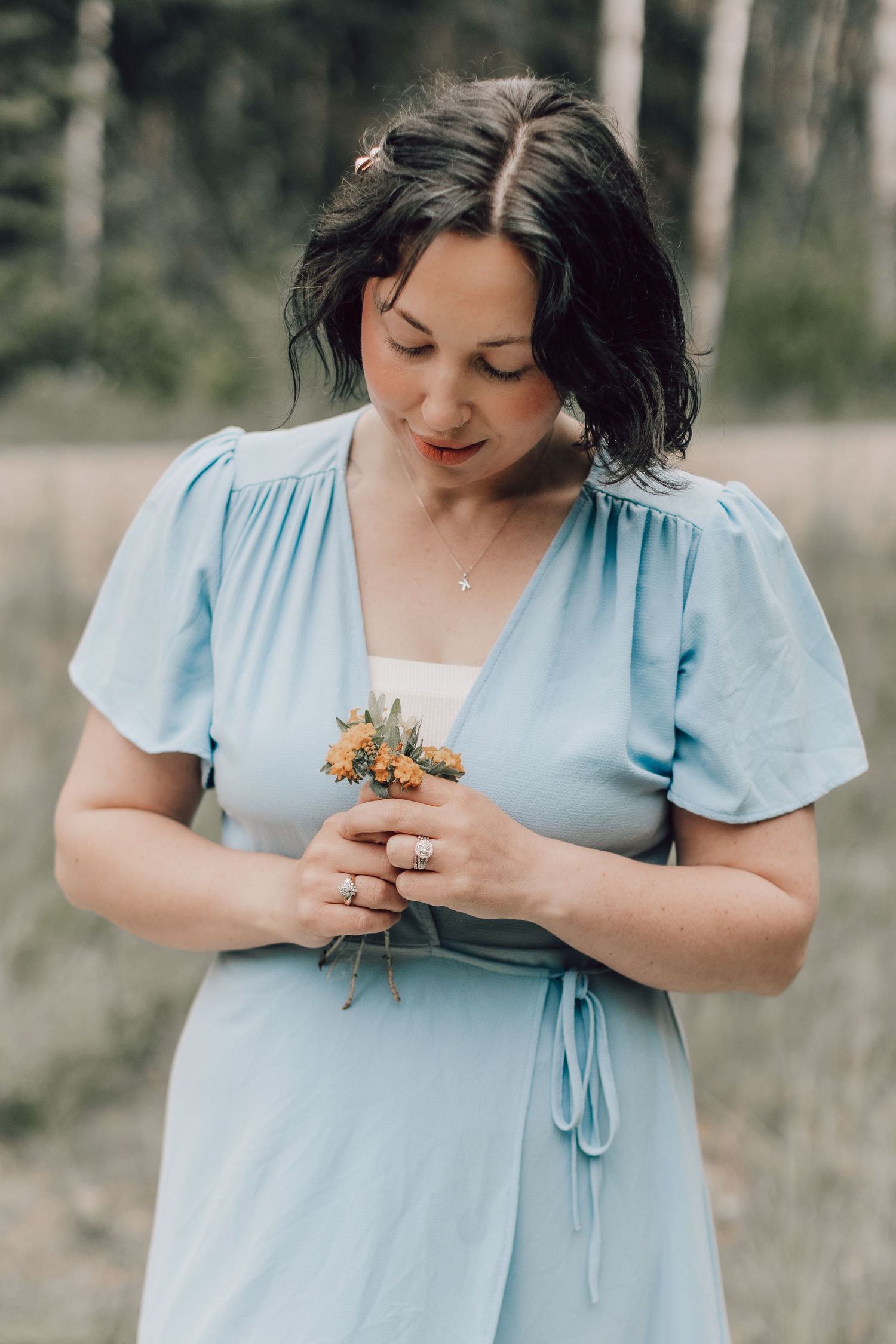 A woman in a blue dress holding a small bouquet of wildflowers while looking down softly.