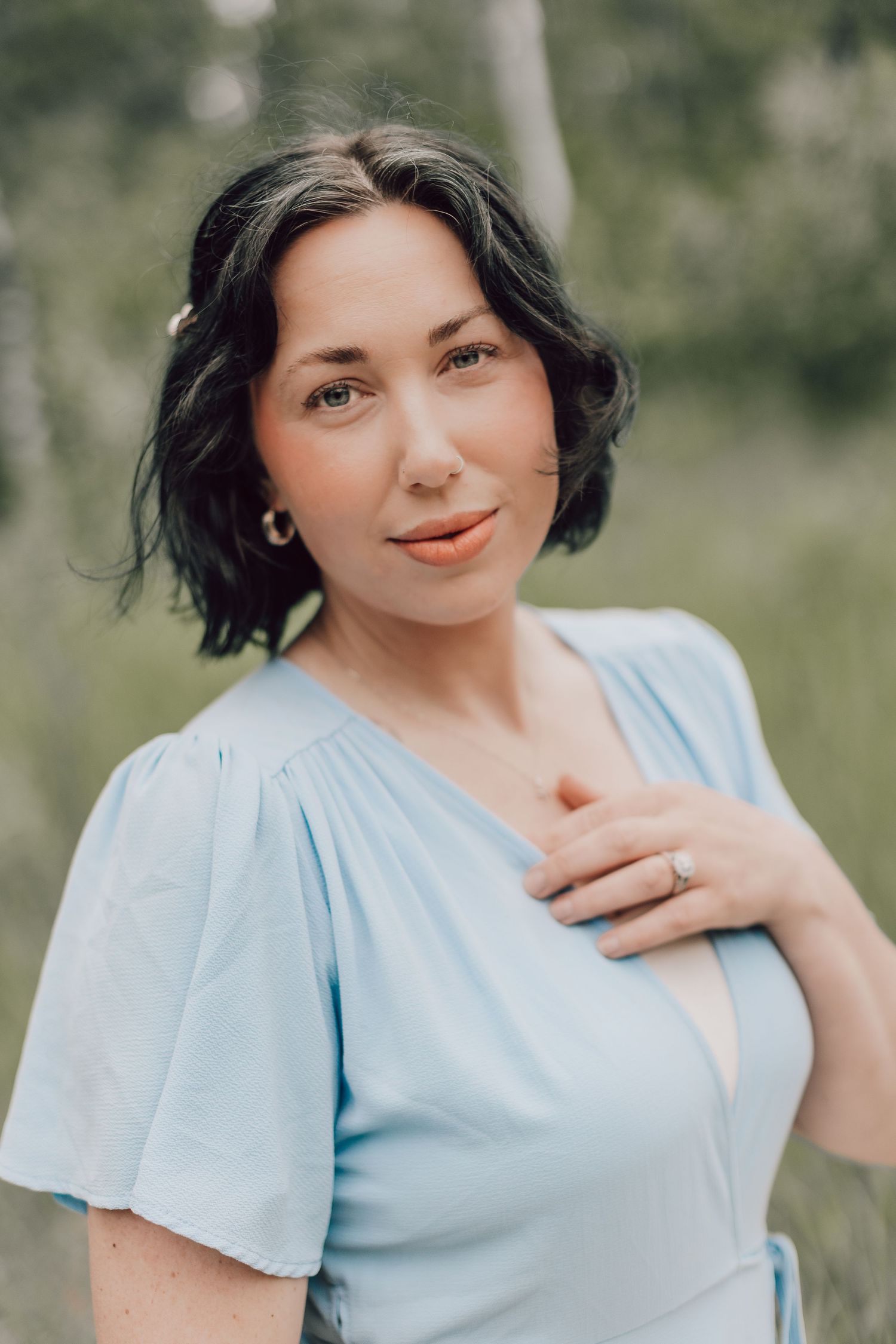 A brunette woman in a light blue dress poses outdoors, gently touching her chest while wearing a ring.
