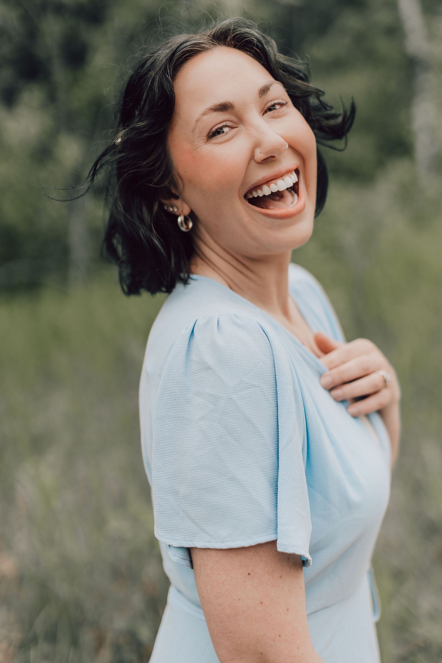 A dark-haired woman in a light blue dress laughs joyfully outdoors in a natural setting.