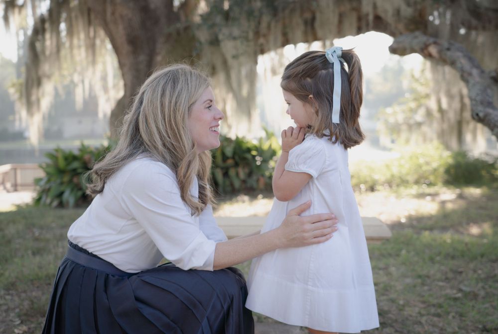 Tender moment between woman and young girl in white dress outdoors with Spanish moss in background.