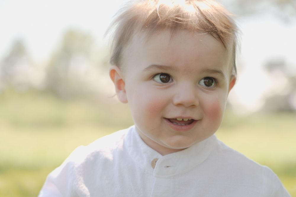 Baby wearing white outfit smiling brightly in outdoor portrait session.