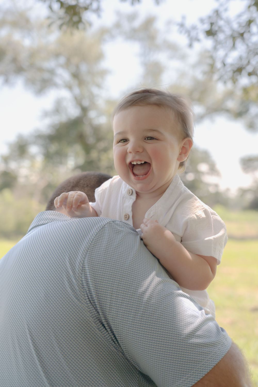 Joyful baby laughing while being held up against sunny outdoor background.