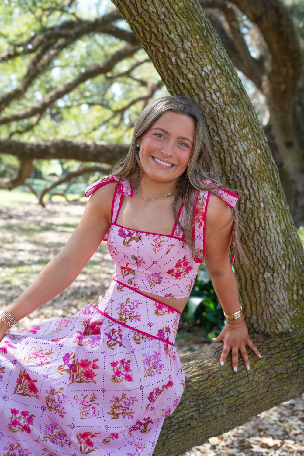A person wearing a pink floral sundress poses playfully near a large tree trunk in a natural outdoor setting.