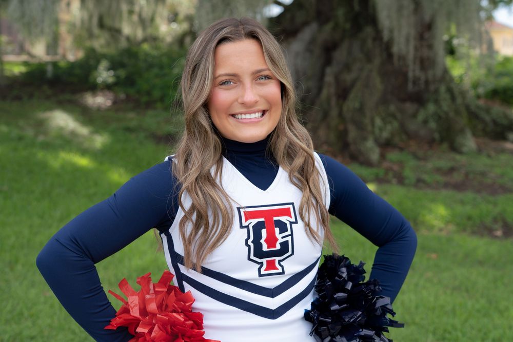 A cheerleader in a navy and white uniform with pom poms poses outdoors on a grassy lawn.