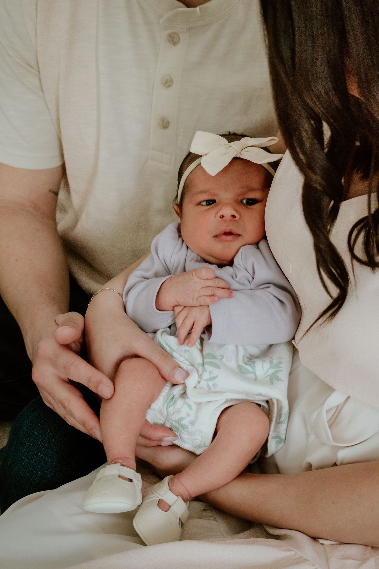 Newborn baby girl in lavender outfit and white bow headband held by parents during family photo session.